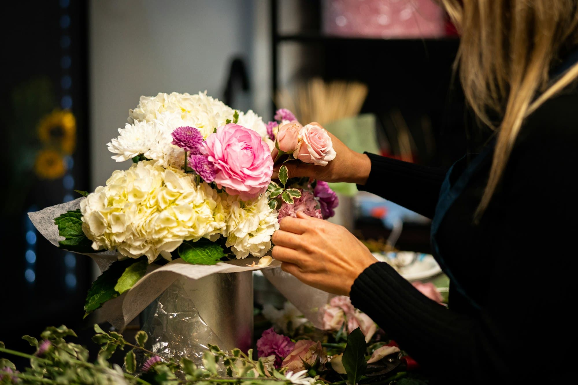 A florist arranging a bouquet of pink roses, white hydrangeas, and chrysanthemums on the workbench.