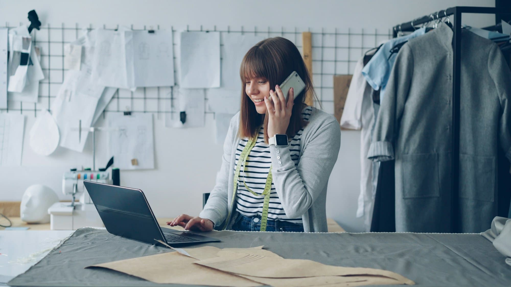A small business owner smiling as she takes a phone call at her workbench, laptop open in front of her.