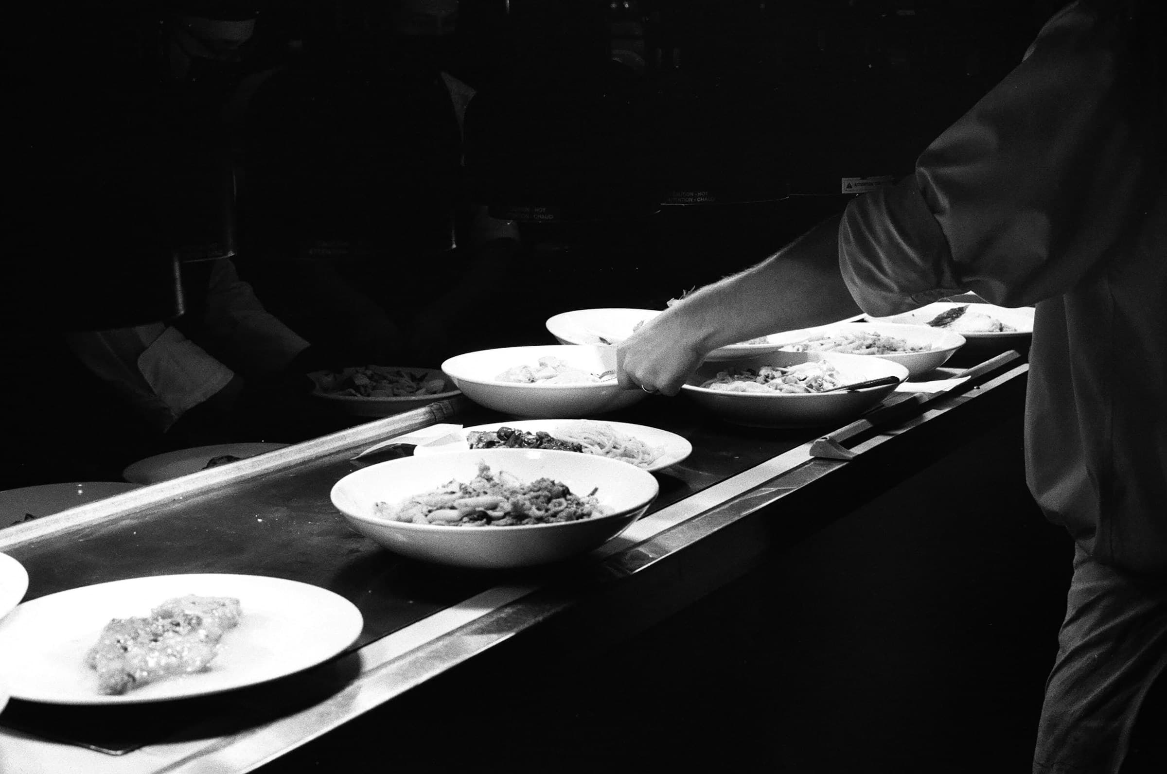 A chef plating a dish on a service line in a dimly lit professional restaurant kitchen.